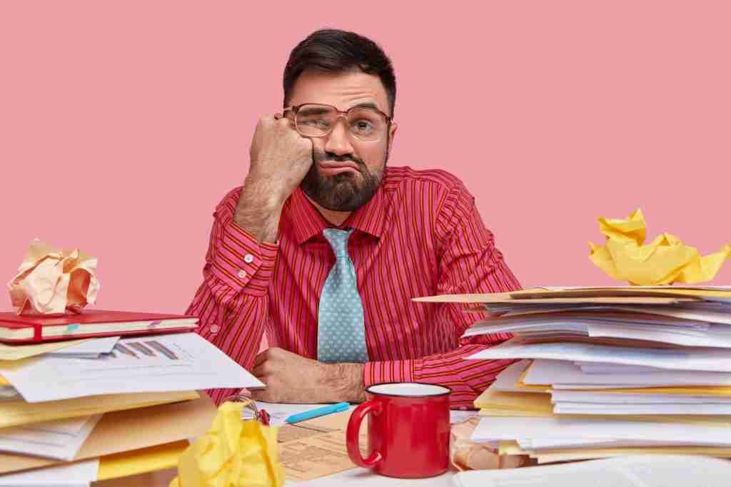 lazy displeased fatigue male worker keeps fist cheek has sleepy look dressed formal shirt tie works with papers has mess workpace