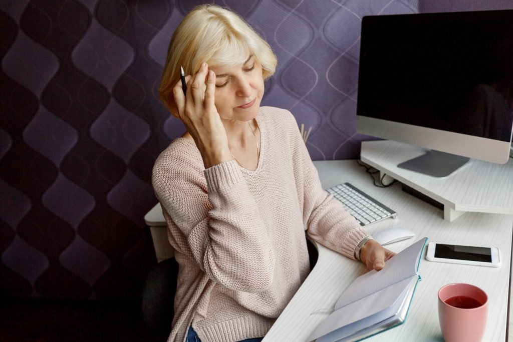 What Are The Effects of Stress On Mental Health? 1 blond mature woman writing her planner while working by computer home