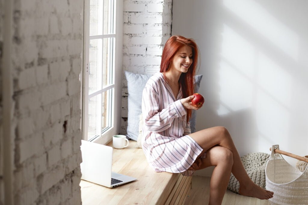 How to Create a Morning Routine That Supports Your Manifestation Practice 5 People, healthy lifestyle, food, diet and morning routine concept. Beautiful red haired young Caucasian woman in striped night suit laughing, enjoying calm peaceful morning at home, eating fresh apple