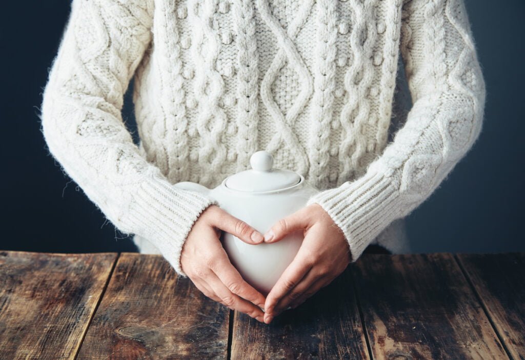 The Role of Forgiveness in Clearing Blockages to Manifestation 2 Woman in warm sweater holds hands on big white teapot with tea in heart shape. Front view, grunge wooden table. anfas, no face.
