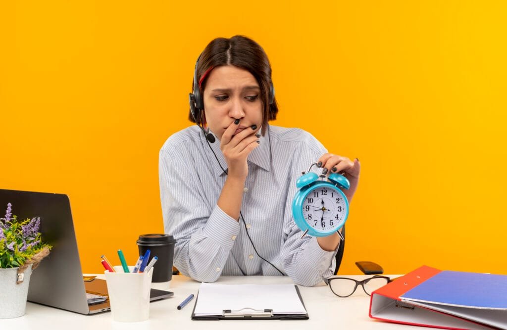 Using the Law of Attraction to Overcome Procrastination and Build Discipline 3 unpleased young call center girl wearing headset sitting at desk with work tools putting hand on mouth holding and looking at alarm clock isolated on orange background